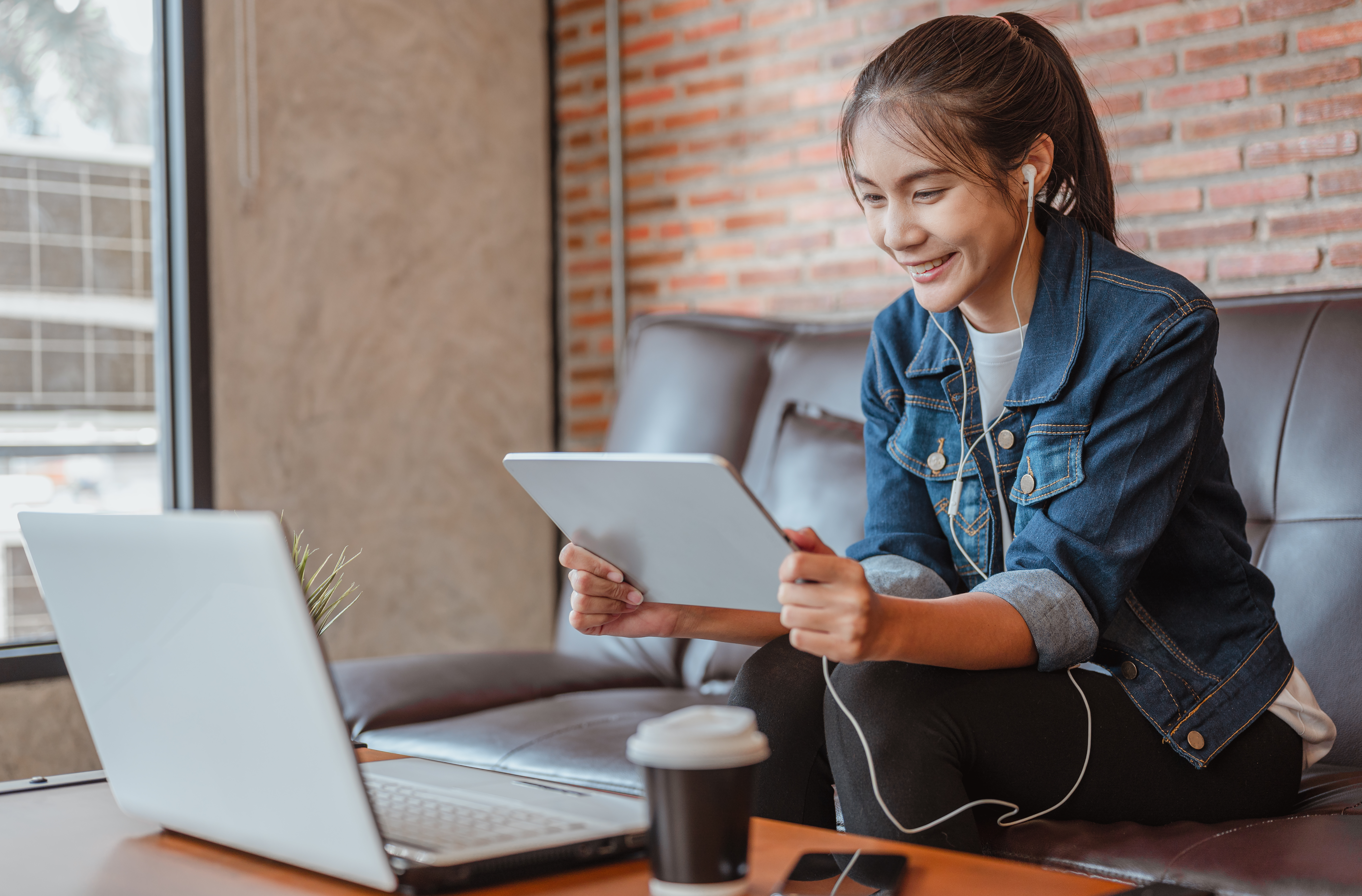 A smiling young woman wearing a denim jacket and earbuds, sitting on a couch and holding a tablet while looking at a laptop on the table.