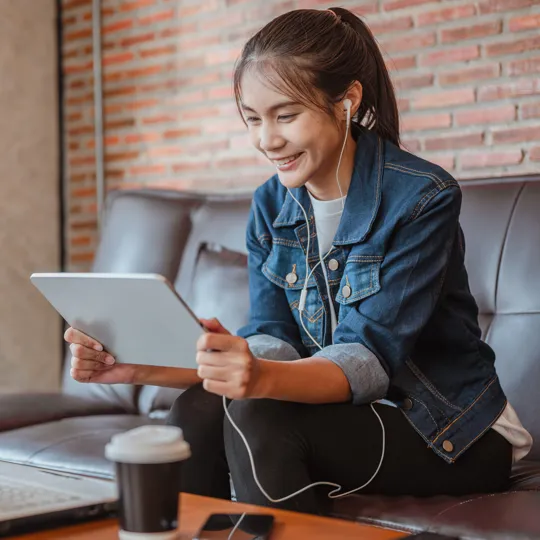 A smiling young woman wearing a denim jacket and earbuds, sitting on a couch and holding a tablet while looking at a laptop on the table.