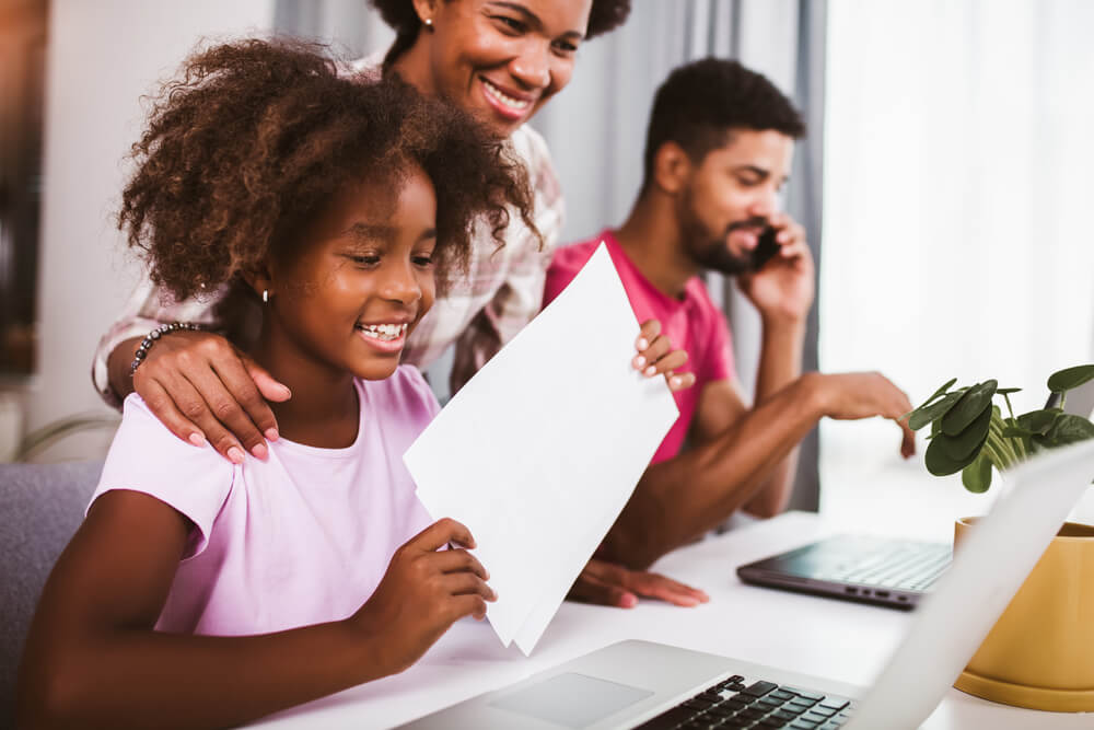 Young child smiling at laptop whilst holding a piece of paper.
