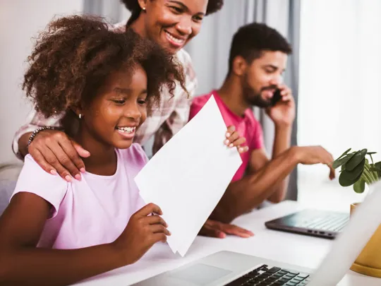 Young child smiling at laptop whilst holding a piece of paper.