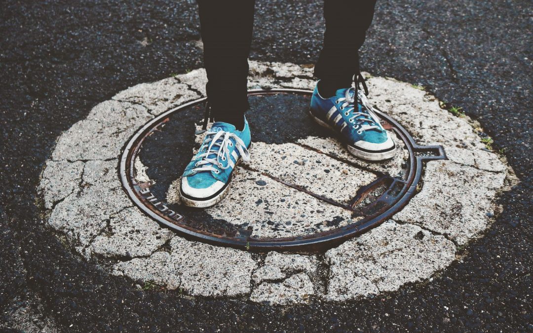 A low-angle shot showing a person's legs and feet standing on a manhole cover in the middle of a street. The person is wearing black trousers and a pair of blue sneakers with white laces. The manhole cover is surrounded by cracked concrete and asphalt.