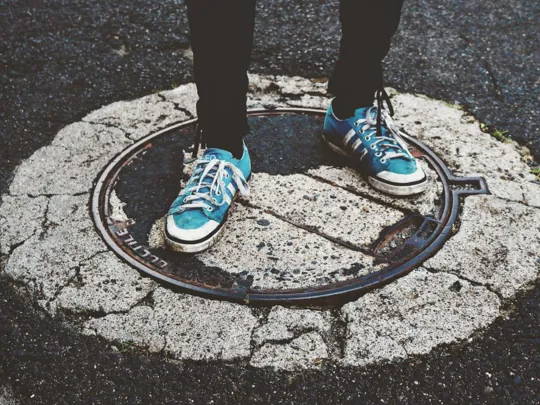 A low-angle shot showing a person's legs and feet standing on a manhole cover in the middle of a street. The person is wearing black trousers and a pair of blue sneakers with white laces. The manhole cover is surrounded by cracked concrete and asphalt.