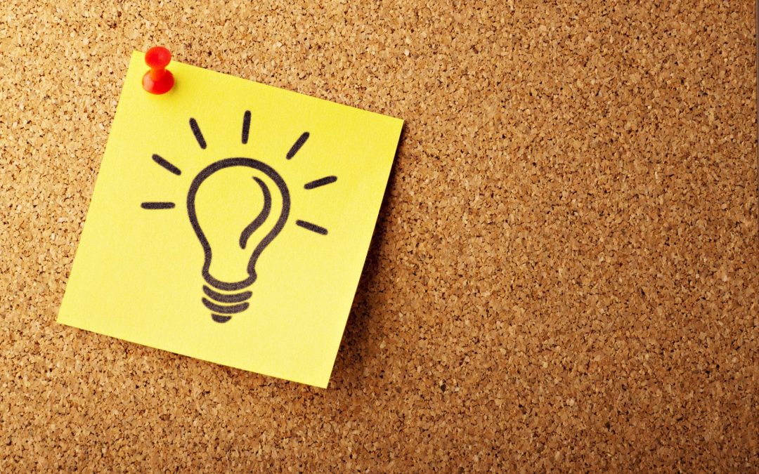 A close-up shot of a yellow sticky note pinned with a red pushpin to a cork board. A black outline drawing of a lightbulb is on the sticky note.