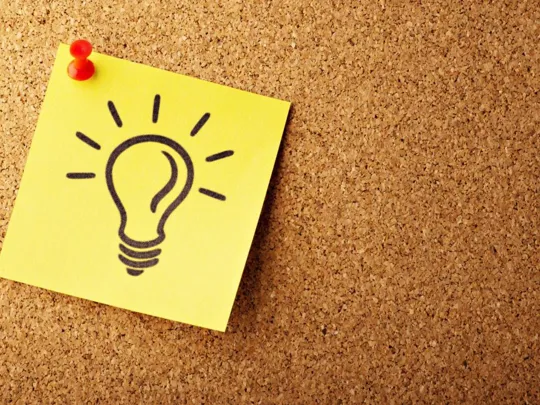 A close-up shot of a yellow sticky note pinned with a red pushpin to a cork board. A black outline drawing of a lightbulb is on the sticky note.
