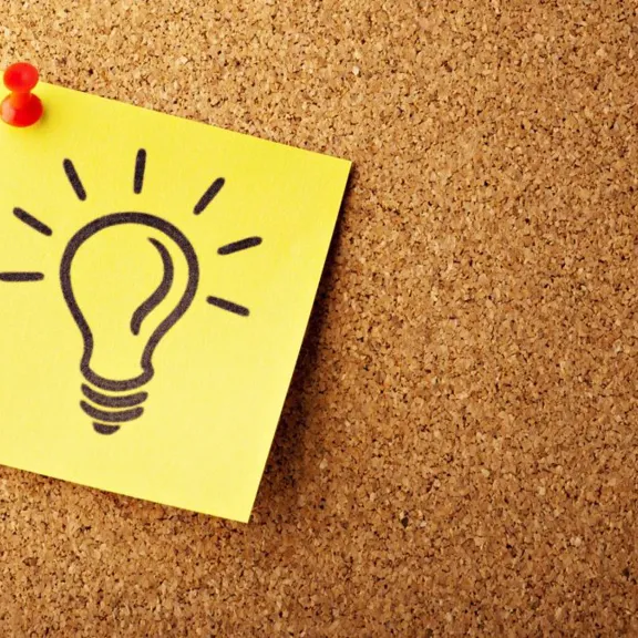 A close-up shot of a yellow sticky note pinned with a red pushpin to a cork board. A black outline drawing of a lightbulb is on the sticky note.