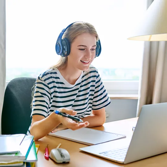 A smiling young woman wearing blue headphones and a striped shirt, holding a pen and looking at a laptop while studying at a desk indoors.