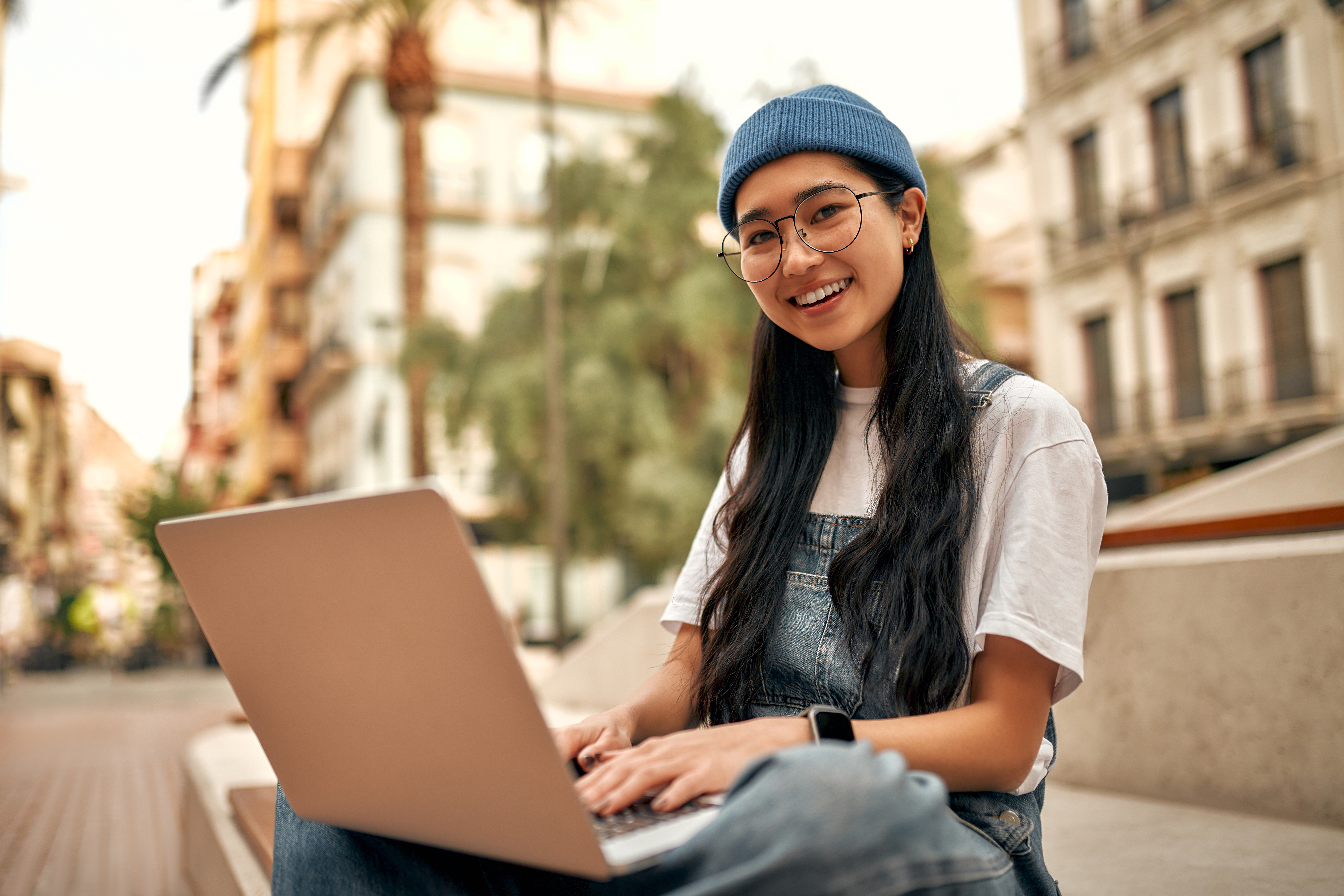 A young woman in a blue beanie and overalls smiles while working on a rose gold laptop outdoors on a sunny city street for online learning