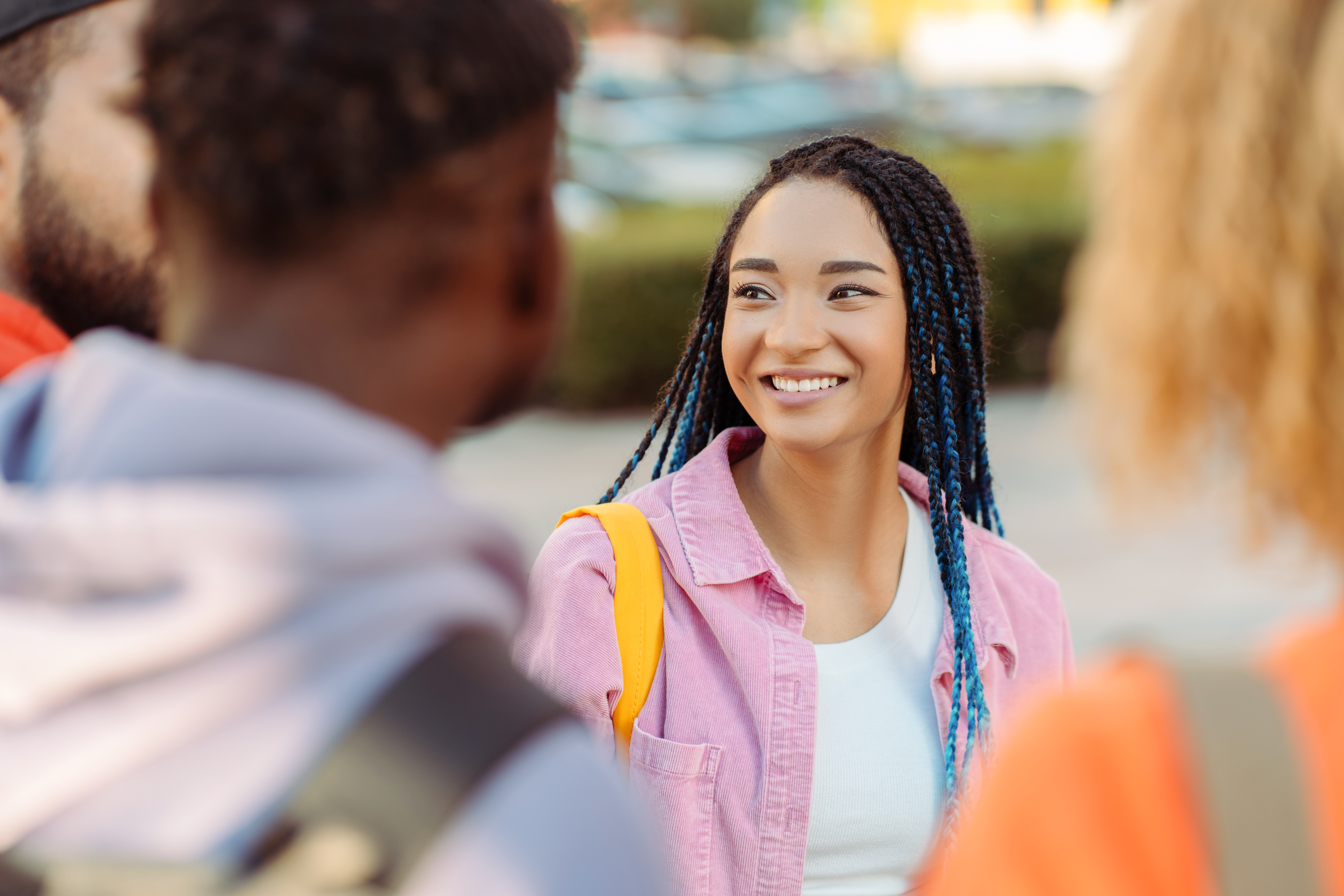A young woman with braided hair and a pink shirt smiles while talking to two blurred classmates outdoors on a sunny school campus.