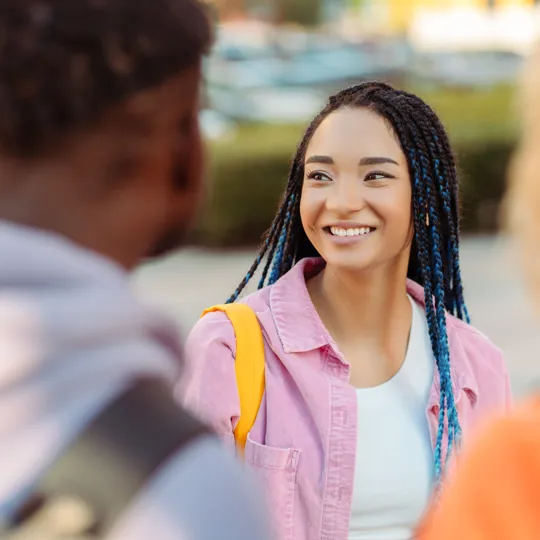 A young woman with braided hair and a pink shirt smiles while talking to two blurred classmates outdoors on a sunny school campus.