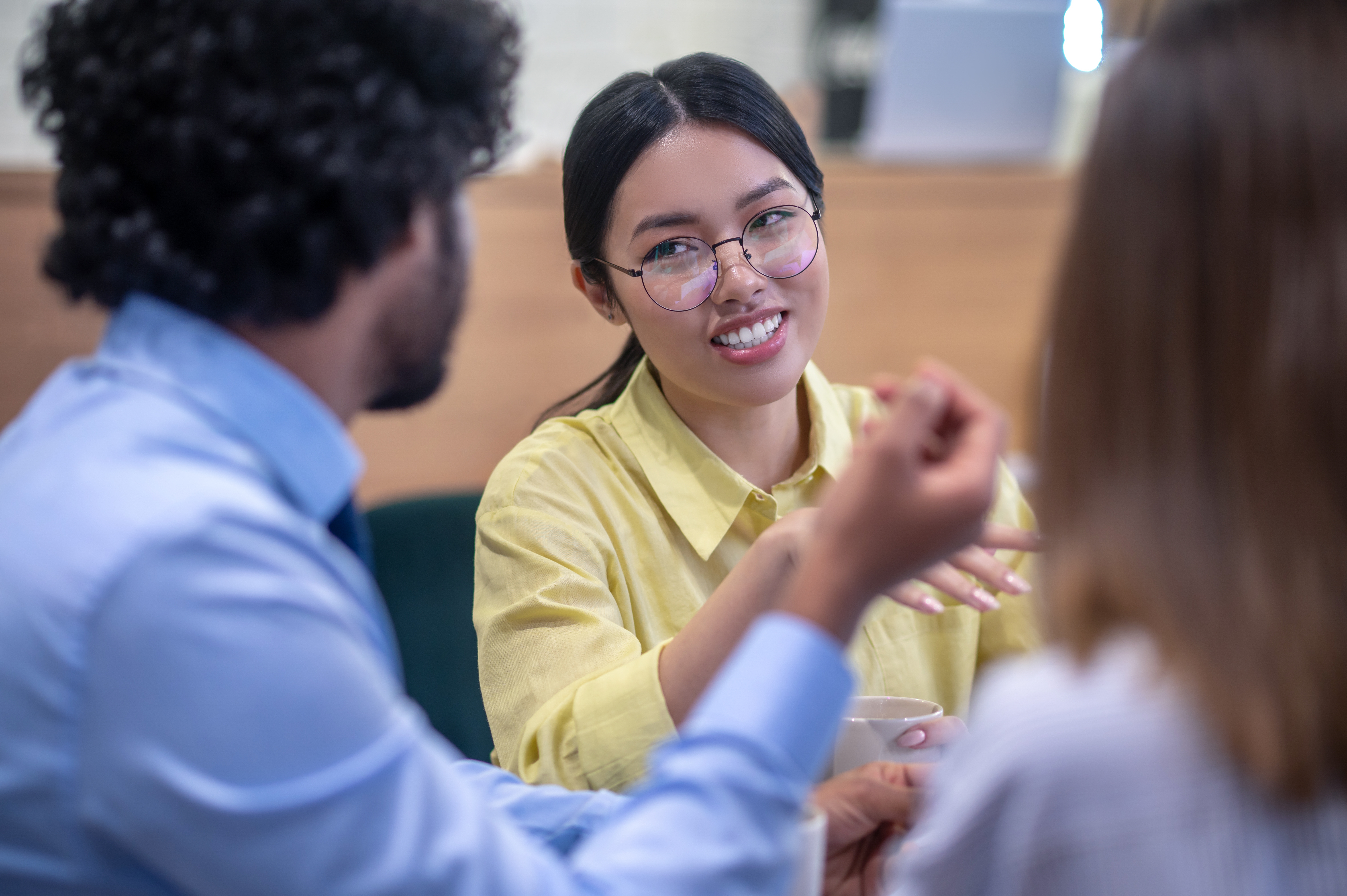 A young woman in a yellow shirt and glasses smiles while gesturing and talking in a small group discussion in a cafe or modern office setting.