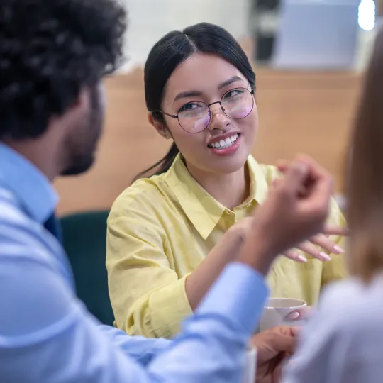 A young woman in a yellow shirt and glasses smiles while gesturing and talking in a small group discussion in a cafe or modern office setting.