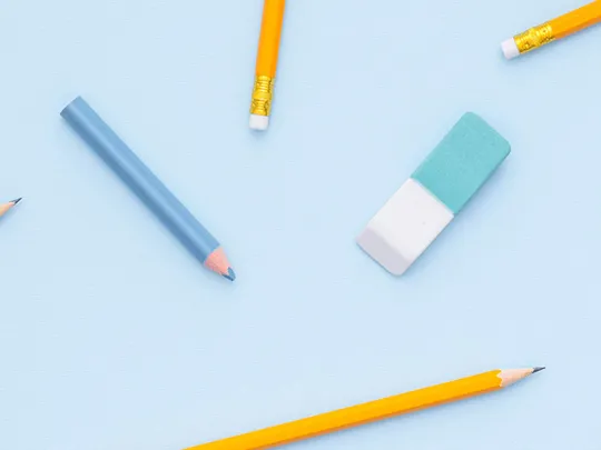 A flat-lay image of various school supplies scattered on a light blue background. The supplies include yellow and colored pencils, a blue eraser, a ruler, and washi tape.