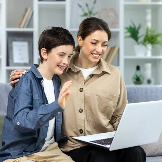 a mother and her son sitting closely on a couch, smiling and waving at a laptop screen during a video call or online class.