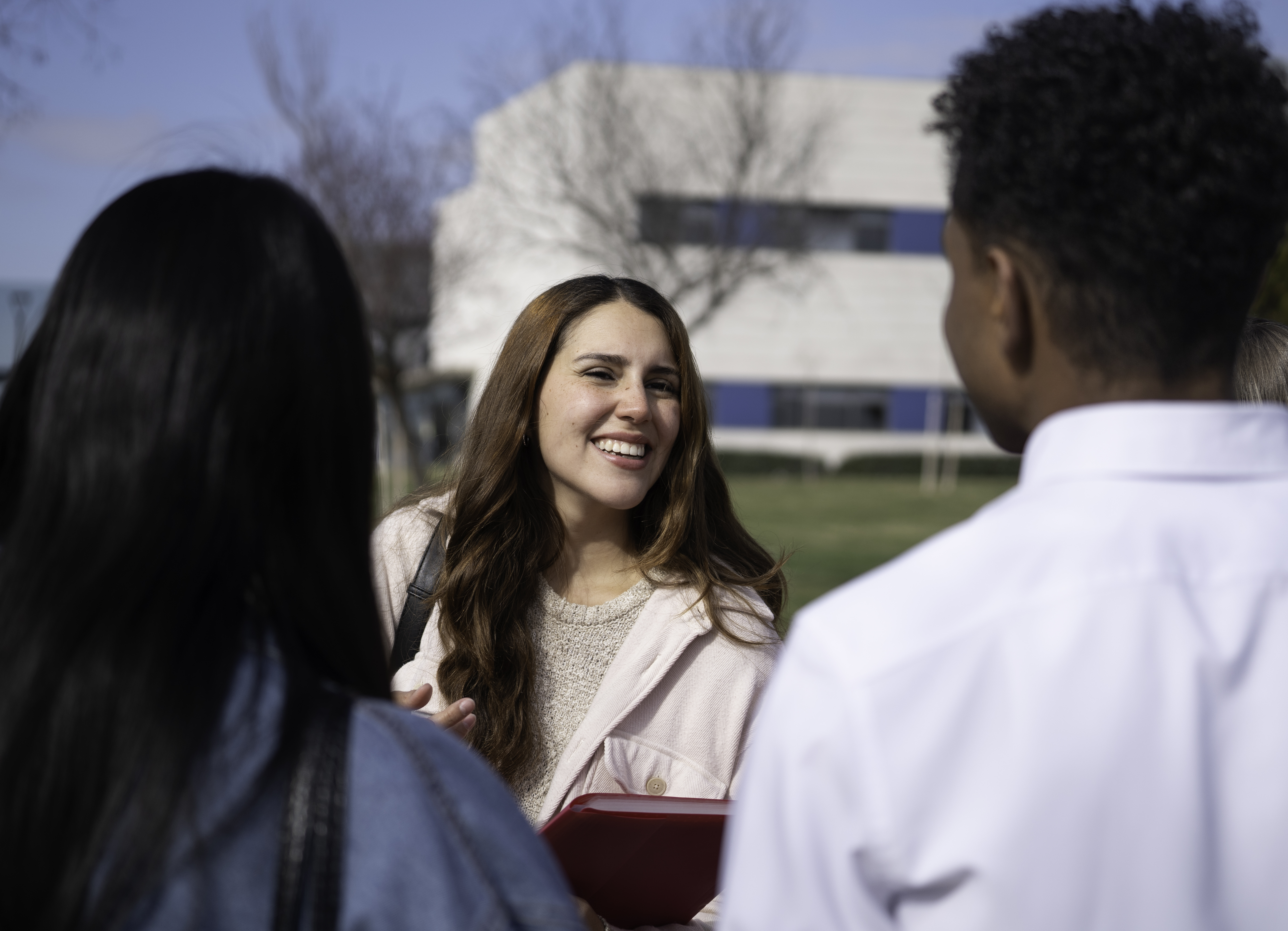 A smiling young woman in a pink jacket holds a red folder while talking with two classmates on a sunny university campus lawn.