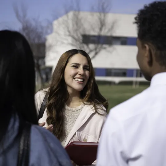 A smiling young woman in a pink jacket holds a red folder while talking with two classmates on a sunny university campus lawn.