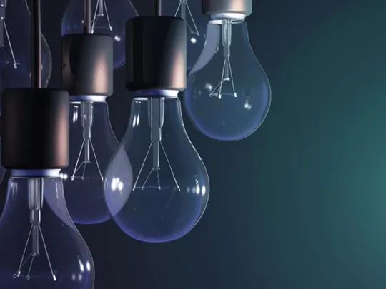 A cluster of unlit, classic filament light bulbs hangs on the left side of the image, while on the right, a single, glowing spiral-shaped fluorescent bulb stands out against a dark background, representing a unique idea or a new way of thinking.