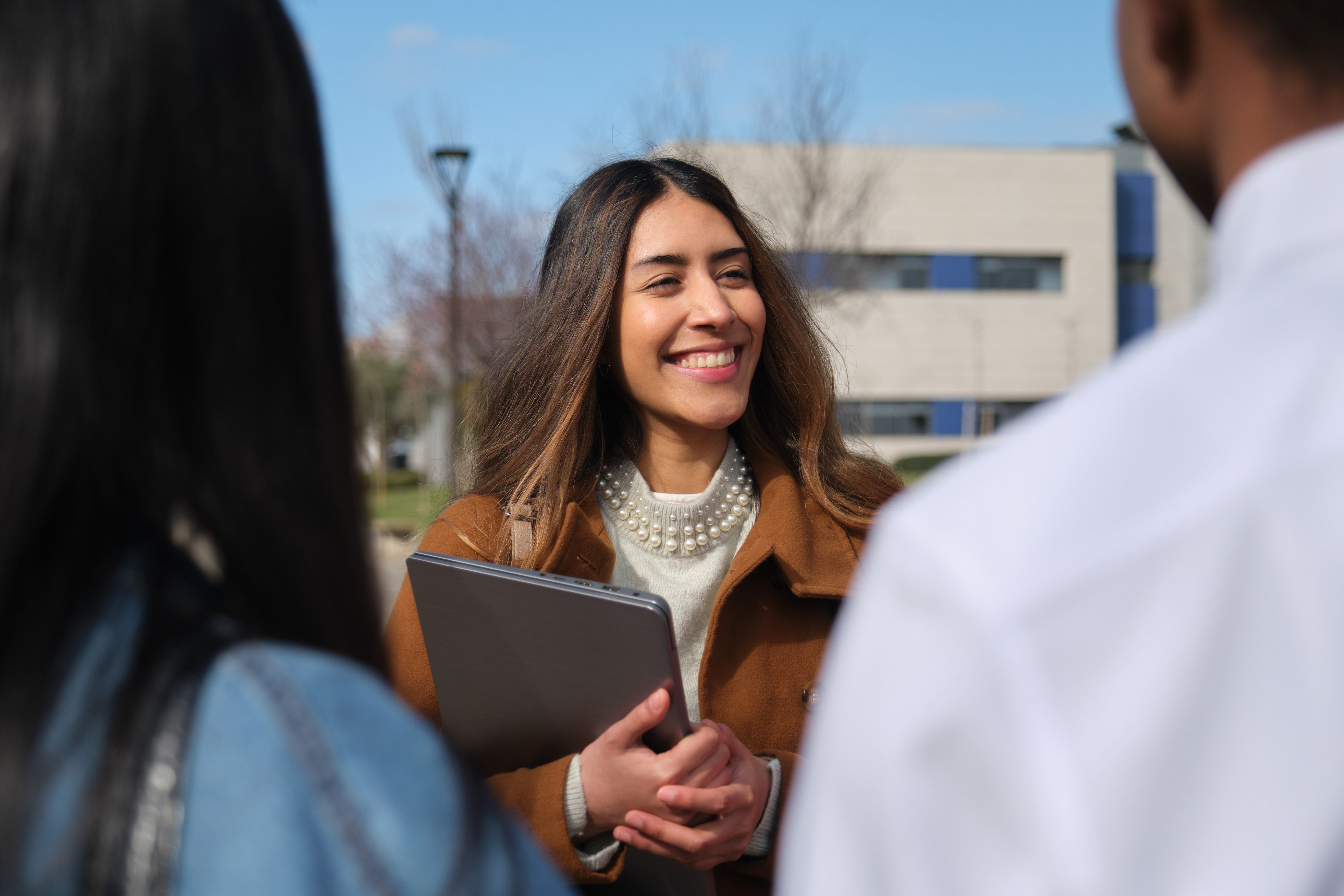 A young woman in a brown coat and pearl necklace smiles while holding a silver laptop, standing outdoors on a sunny university campus.