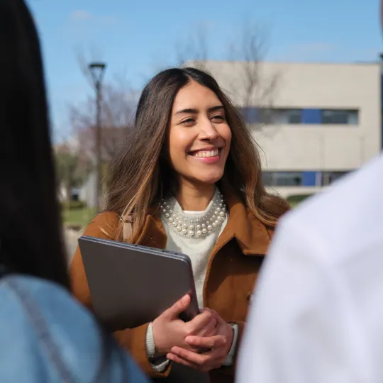 A young woman in a brown coat and pearl necklace smiles while holding a silver laptop, standing outdoors on a sunny university campus.