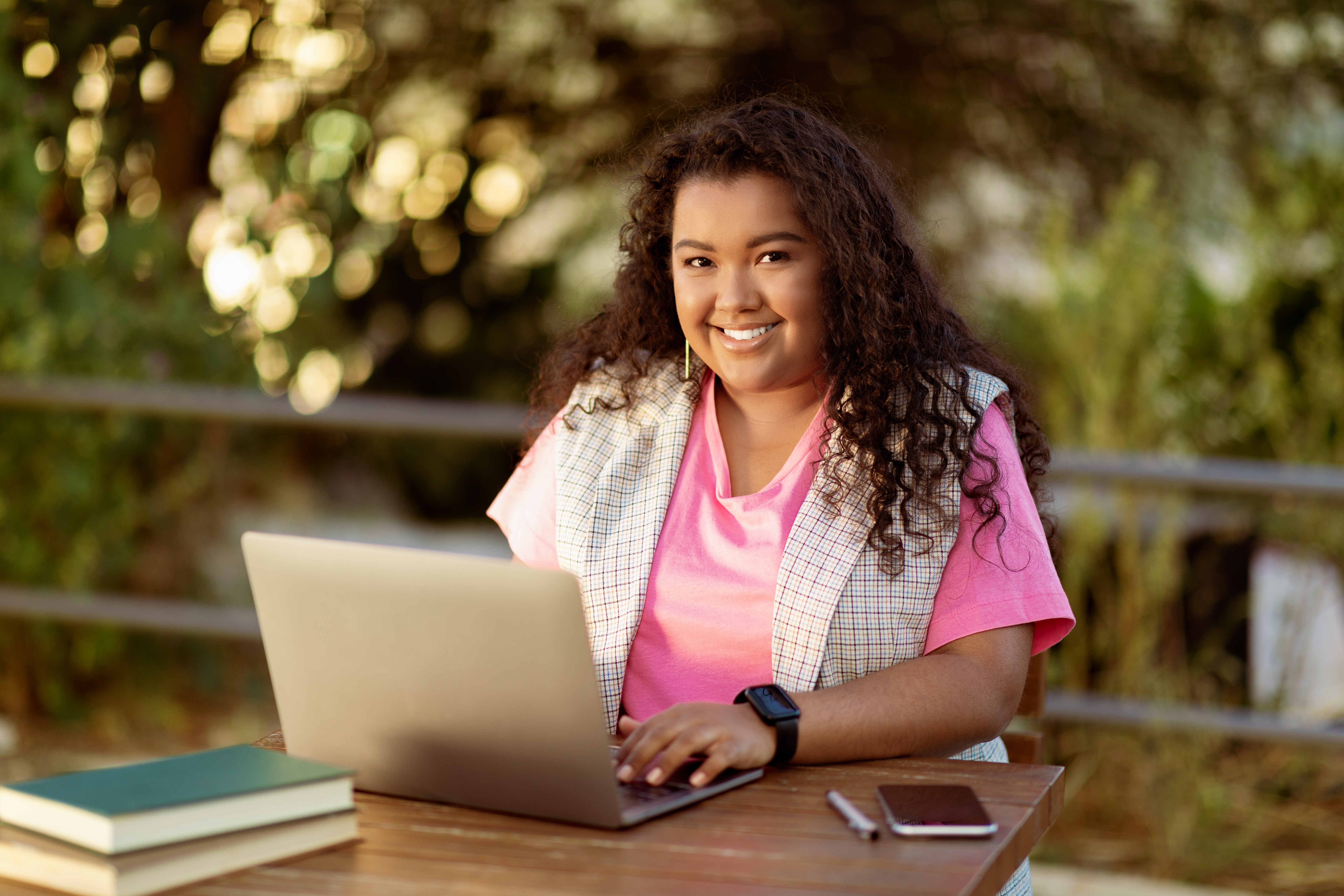 A smiling young student with long, curly dark hair sits outdoors at a wooden table, working on a laptop. She is wearing a bright pink shirt and a light vest. A stack of books, a smartphone, and a pen are on the table next to her, with a blurred natural green background.