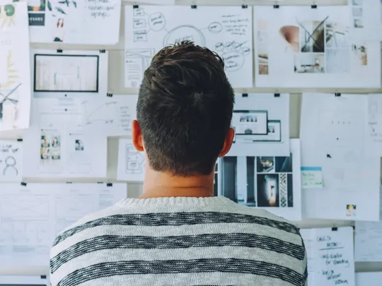 A person is seen from behind, standing in front of a wall covered in papers, sketches, and notes. The person is wearing a striped sweater.