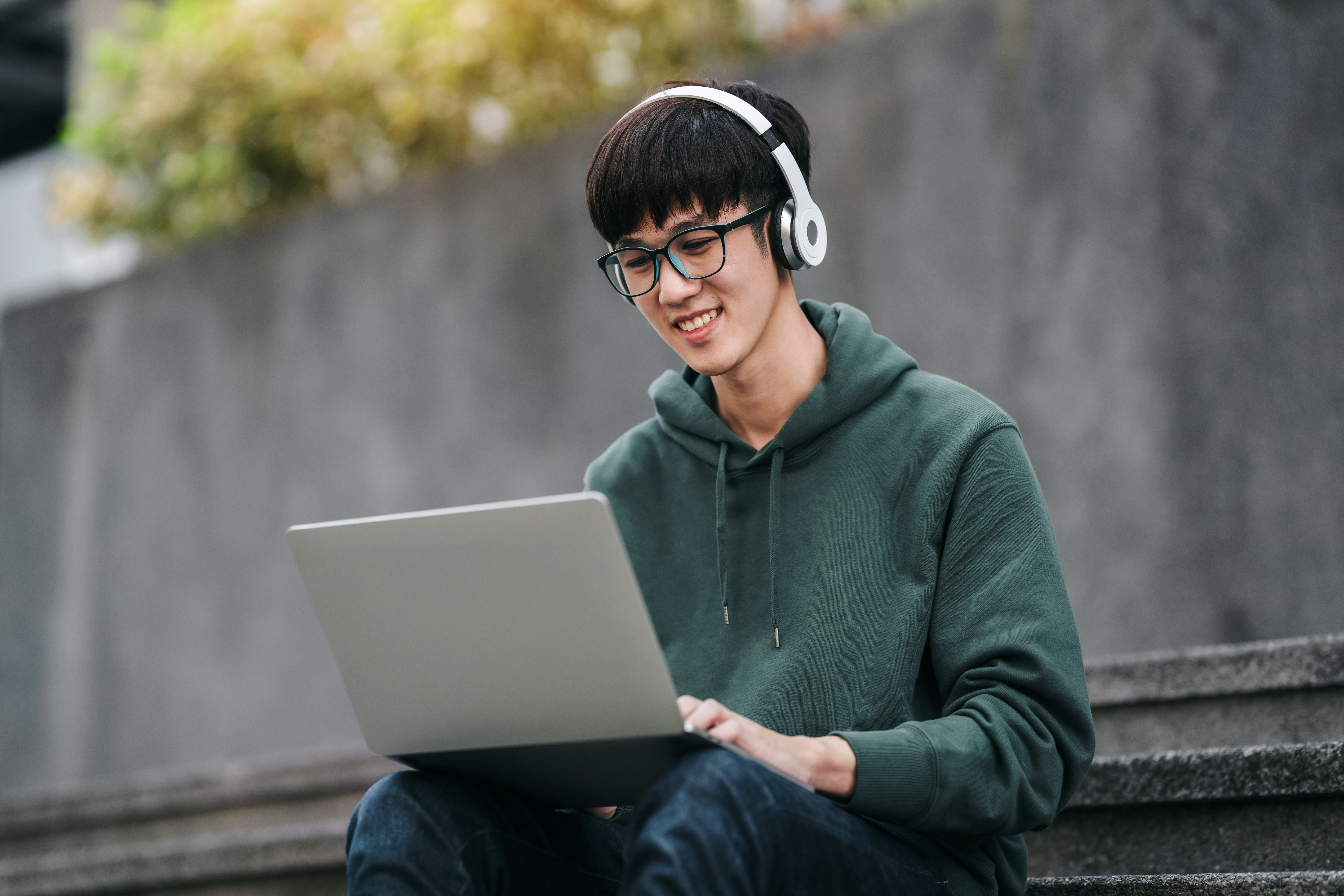 A young male student wearing a green hoodie and white headphones sits on concrete steps, smiling while working on a silver laptop outdoors.