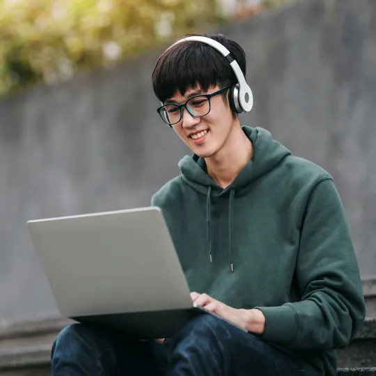A young male student wearing a green hoodie and white headphones sits on concrete steps, smiling while working on a silver laptop outdoors.
