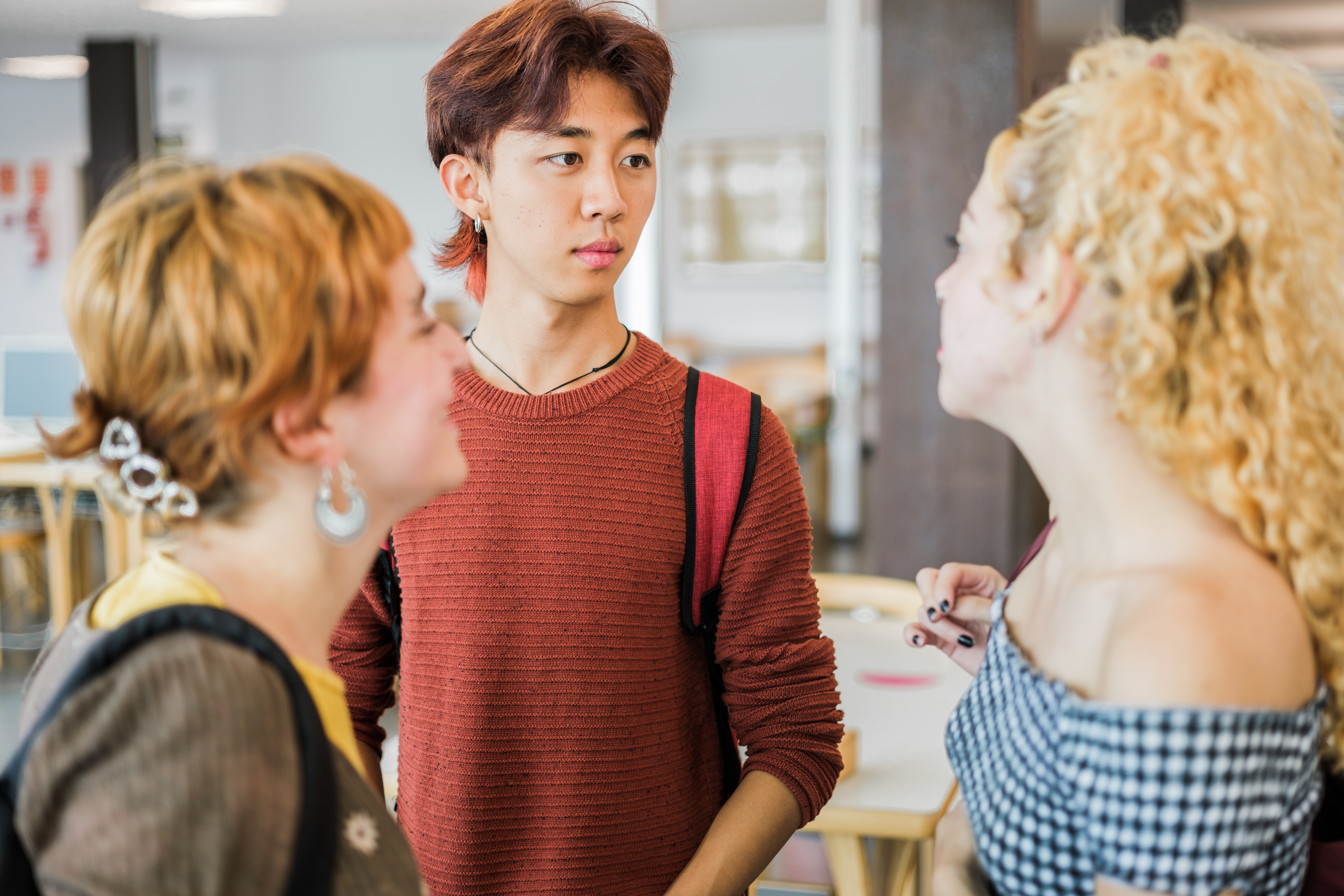 Three young university or high school students are engaged in a conversation indoors, possibly in a classroom or cafe setting. A male student in a terracotta-colored sweater is looking intently at a female student with curly blonde hair. Another female student is visible on the left.