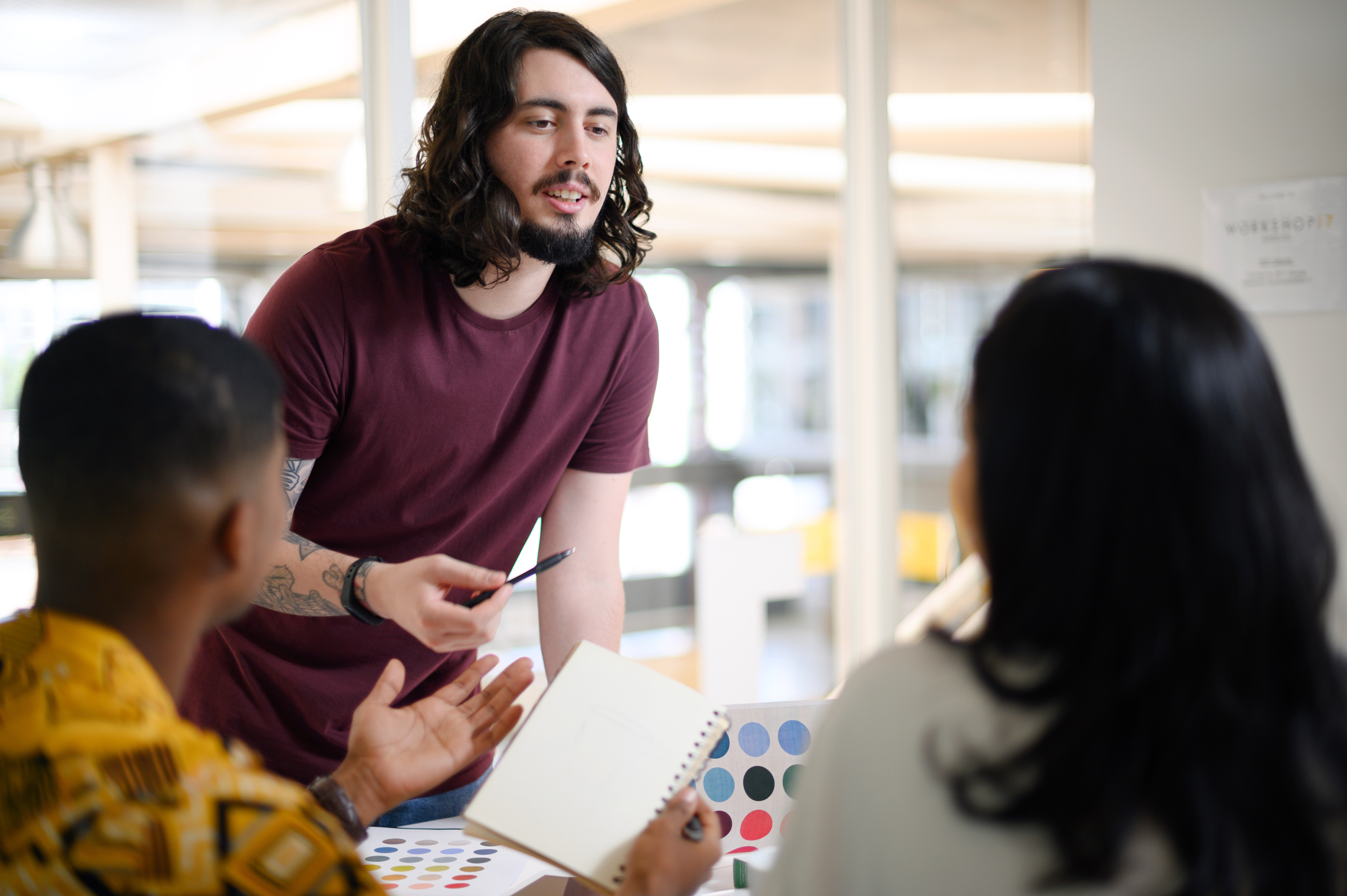 A man with long hair and a maroon shirt stands and talks to two people sitting at a table in a bright, modern office, engaged in an economics discussion.