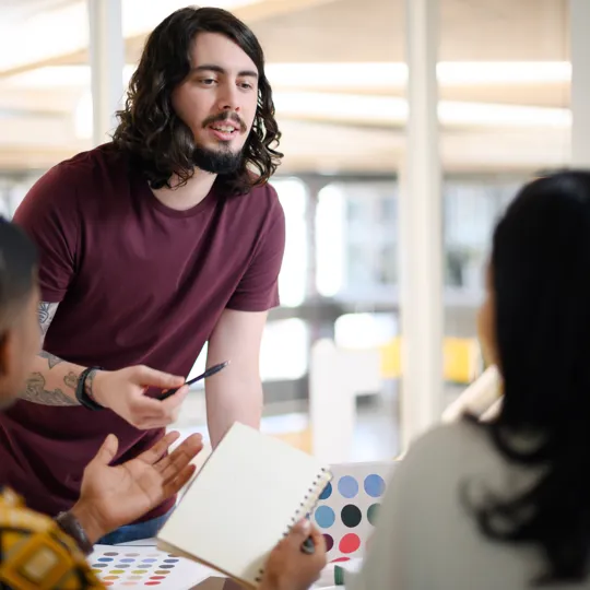 A man with long hair and a maroon shirt stands and talks to two people sitting at a table in a bright, modern office, engaged in an economics discussion.