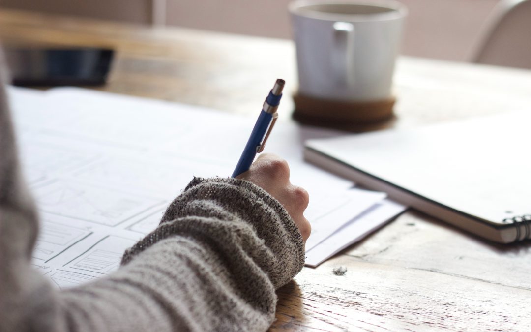 A close-up, over-the-shoulder shot of a person's hand holding a pen and writing on a piece of paper. The person is wearing a gray sweater. A coffee cup and a notebook are on the wooden table.