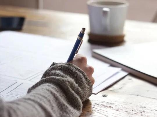 A close-up, over-the-shoulder shot of a person's hand holding a pen and writing on a piece of paper. The person is wearing a gray sweater. A coffee cup and a notebook are on the wooden table.