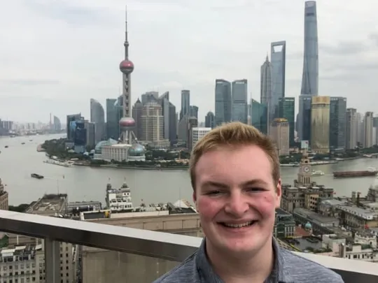 A young man is smiling on a rooftop overlooking the Pudong skyline of Shanghai, China, which includes the Oriental Pearl Tower and the Shanghai World Financial Center, with the Huangpu River flowing in the background.