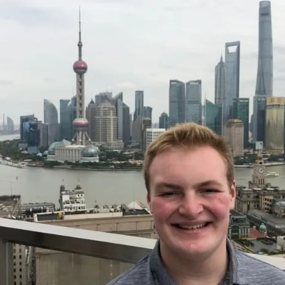 A young man is smiling on a rooftop overlooking the Pudong skyline of Shanghai, China, which includes the Oriental Pearl Tower and the Shanghai World Financial Center, with the Huangpu River flowing in the background.