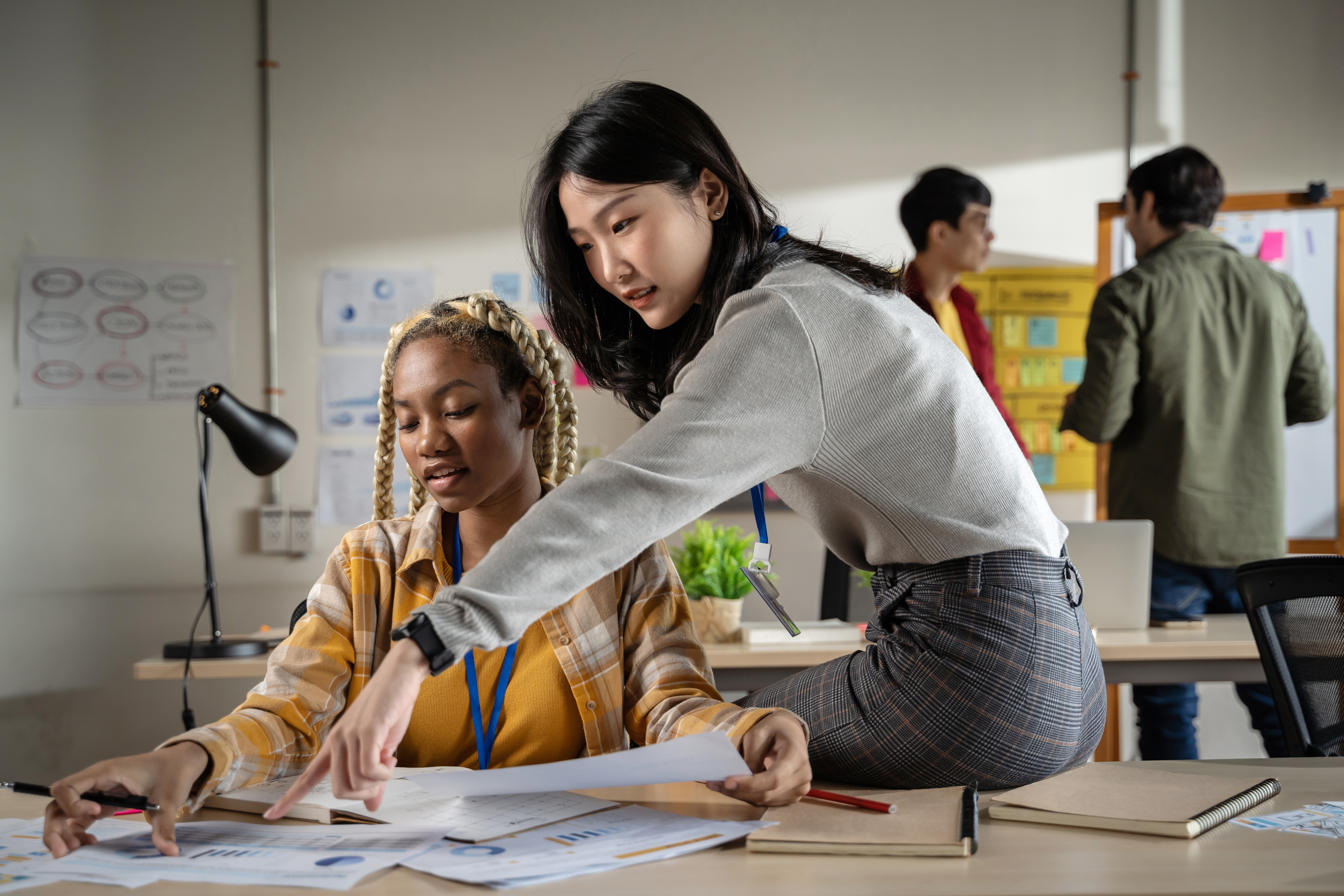 Two women reviewing papers and pointing at a desk in a modern office or classroom while others work in the background.