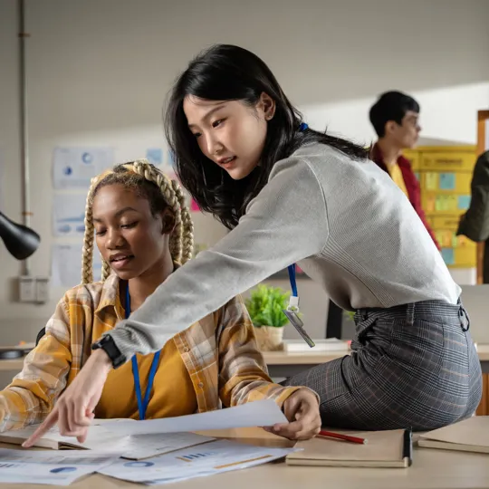Two women reviewing papers and pointing at a desk in a modern office or classroom while others work in the background.