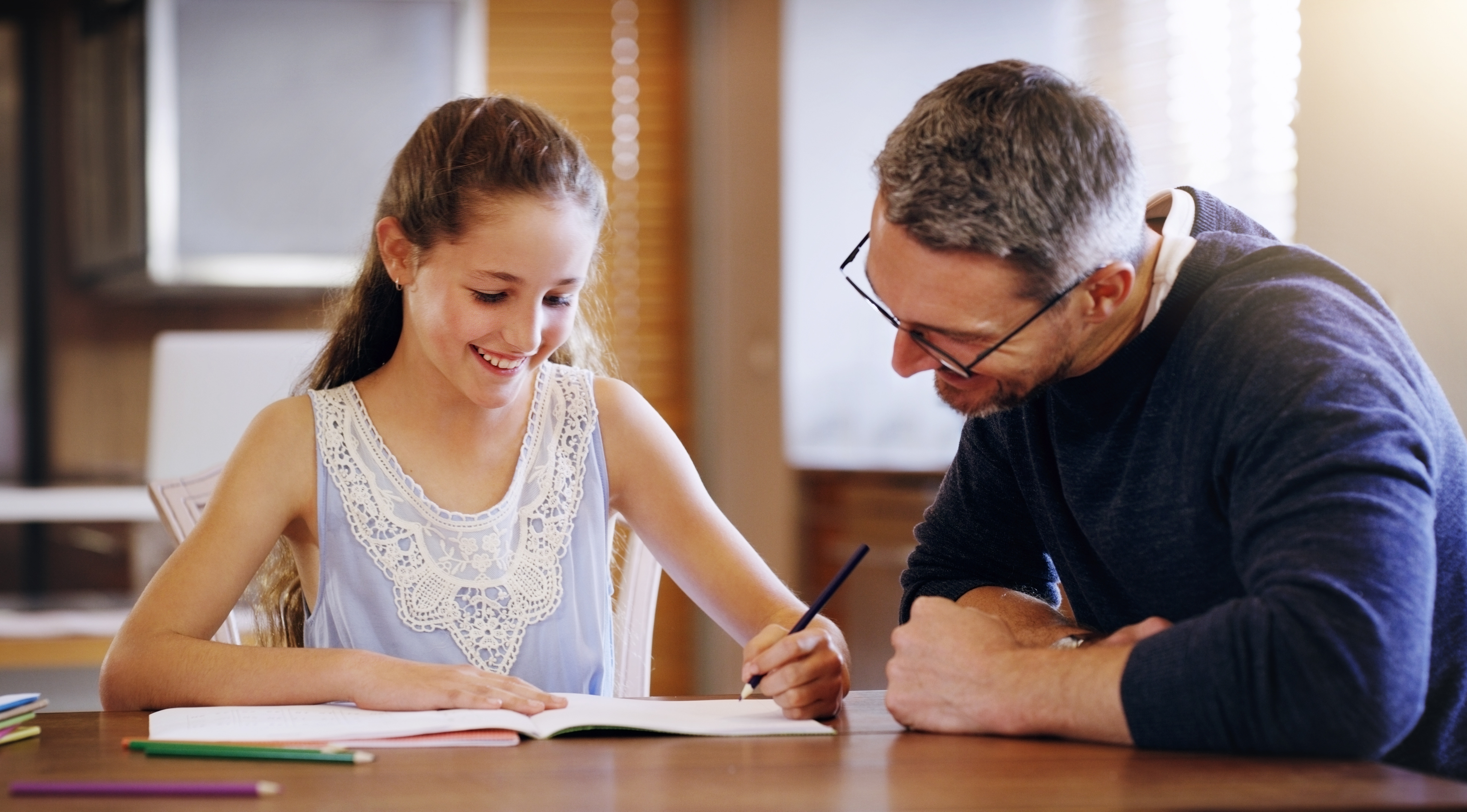 a smiling man and girl sitting together at a table, with the girl writing in a notebook as the man looks on.
