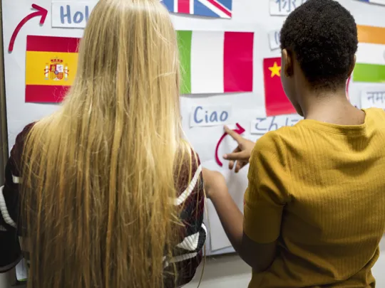Two students pointing at international flags and foreign words (Ciao, Hola, Namaste) on a whiteboard, representing language and cultural studies.