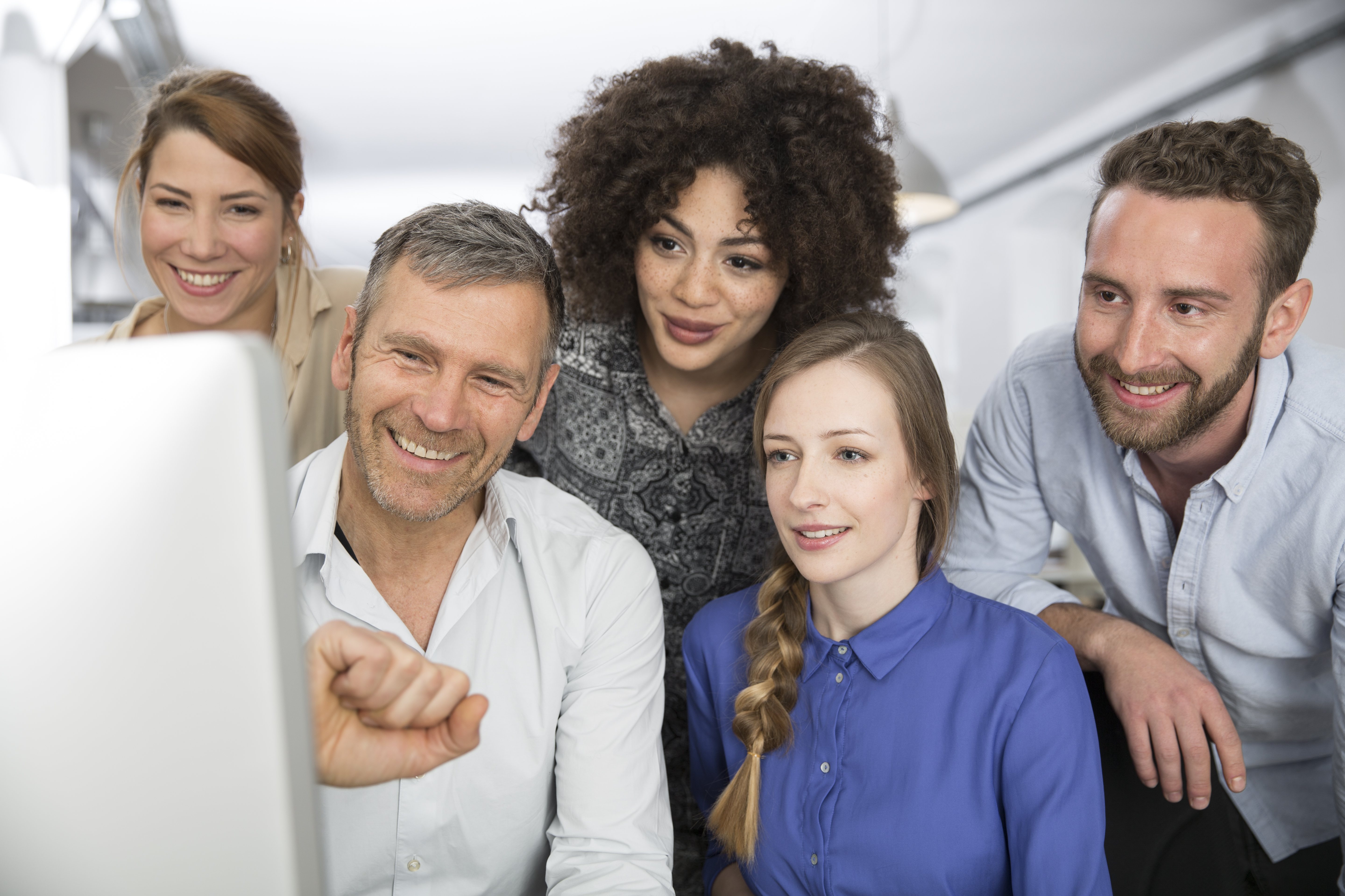 A diverse group of five smiling colleagues or team members are gathered around a computer screen, collaborating on a project in a bright office environment.