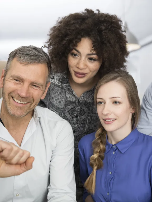 A diverse group of five smiling colleagues or team members are gathered around a computer screen, collaborating on a project in a bright office environment.
