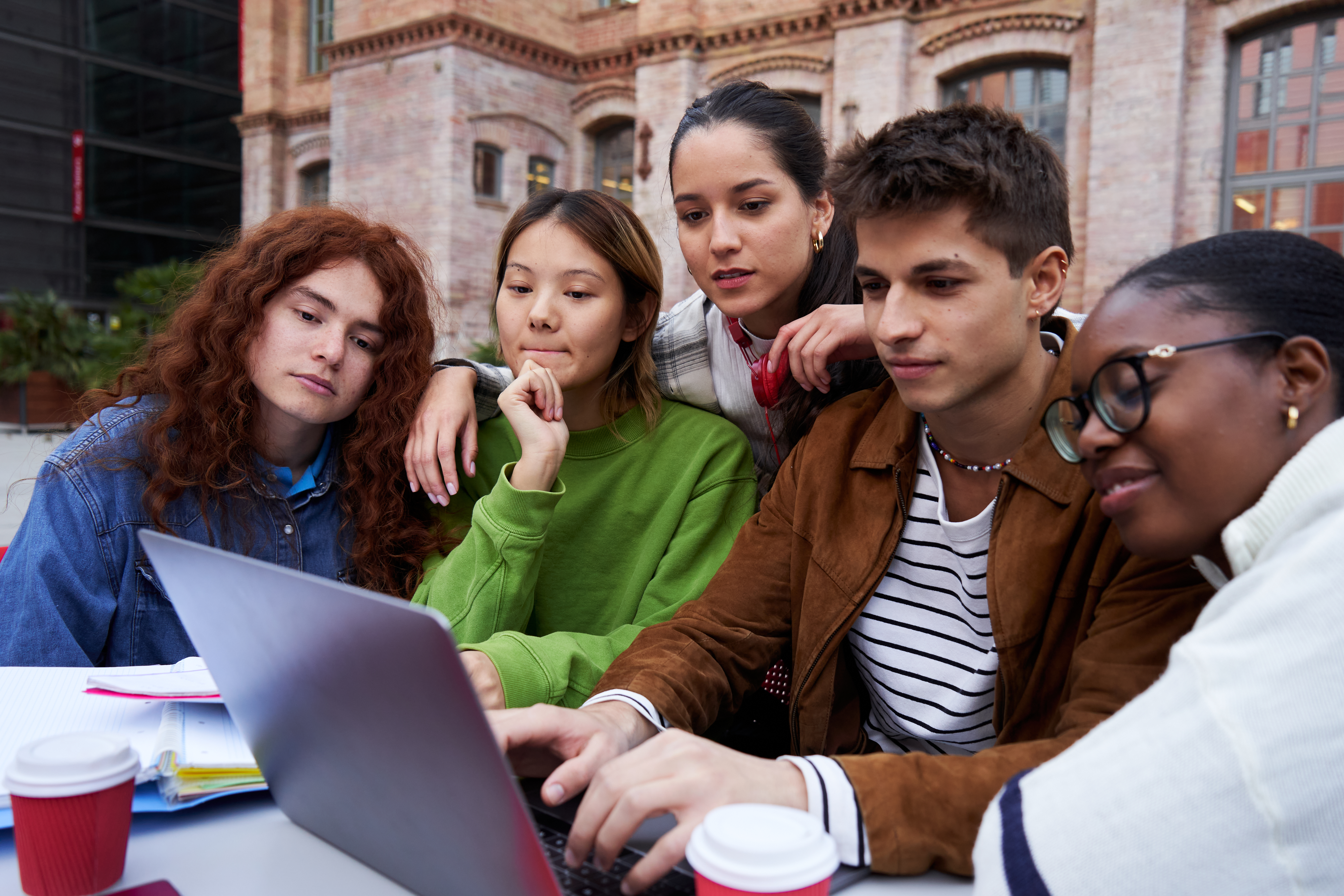 A diverse group of five high school or university students are gathered closely around a laptop, focused on the screen while studying outdoors at a table. They appear engaged in a collaborative project or group study session, with a historic brick building in the background.
