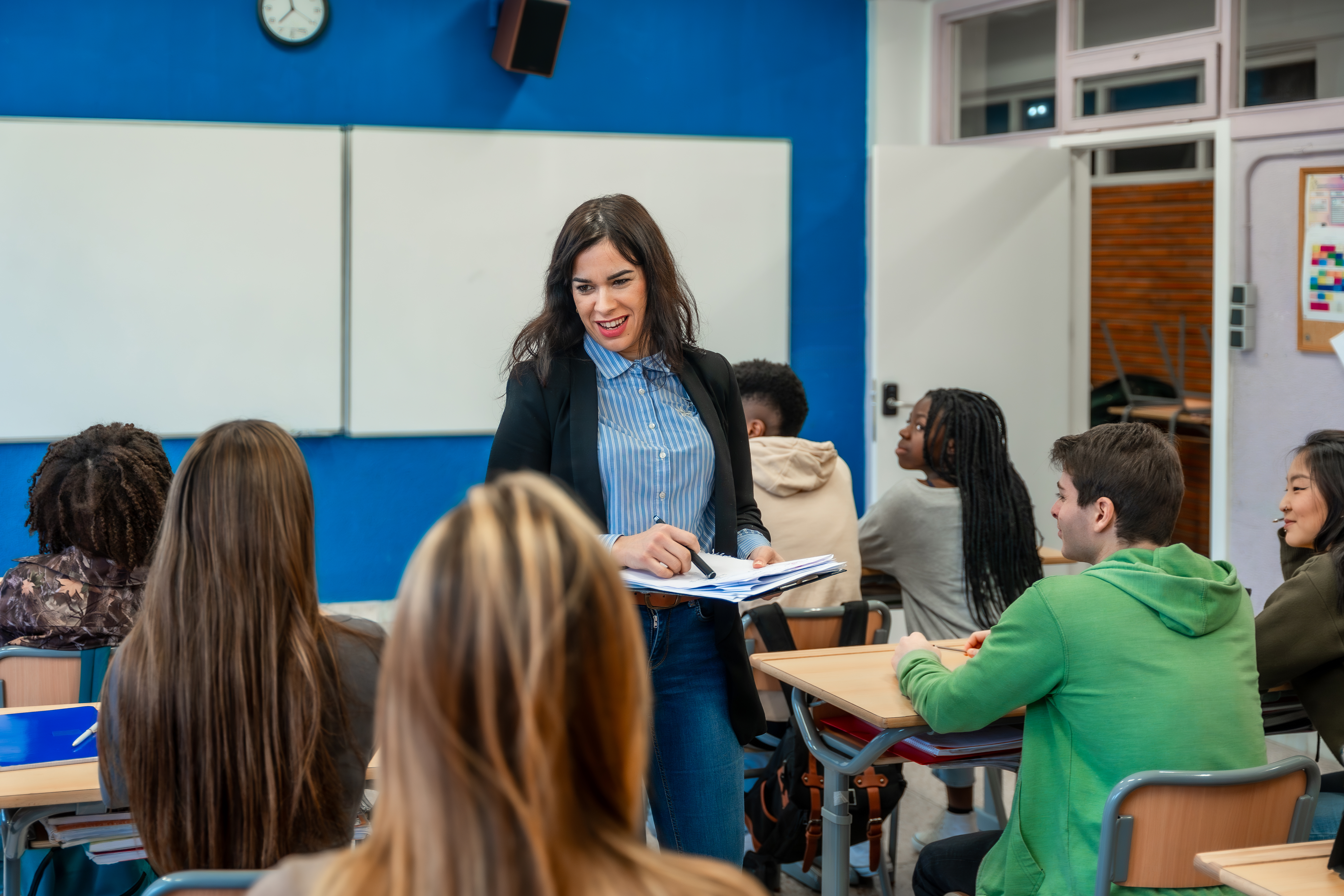 A smiling female teacher in a black blazer stands in the middle of a diverse classroom, addressing students seated at desks.