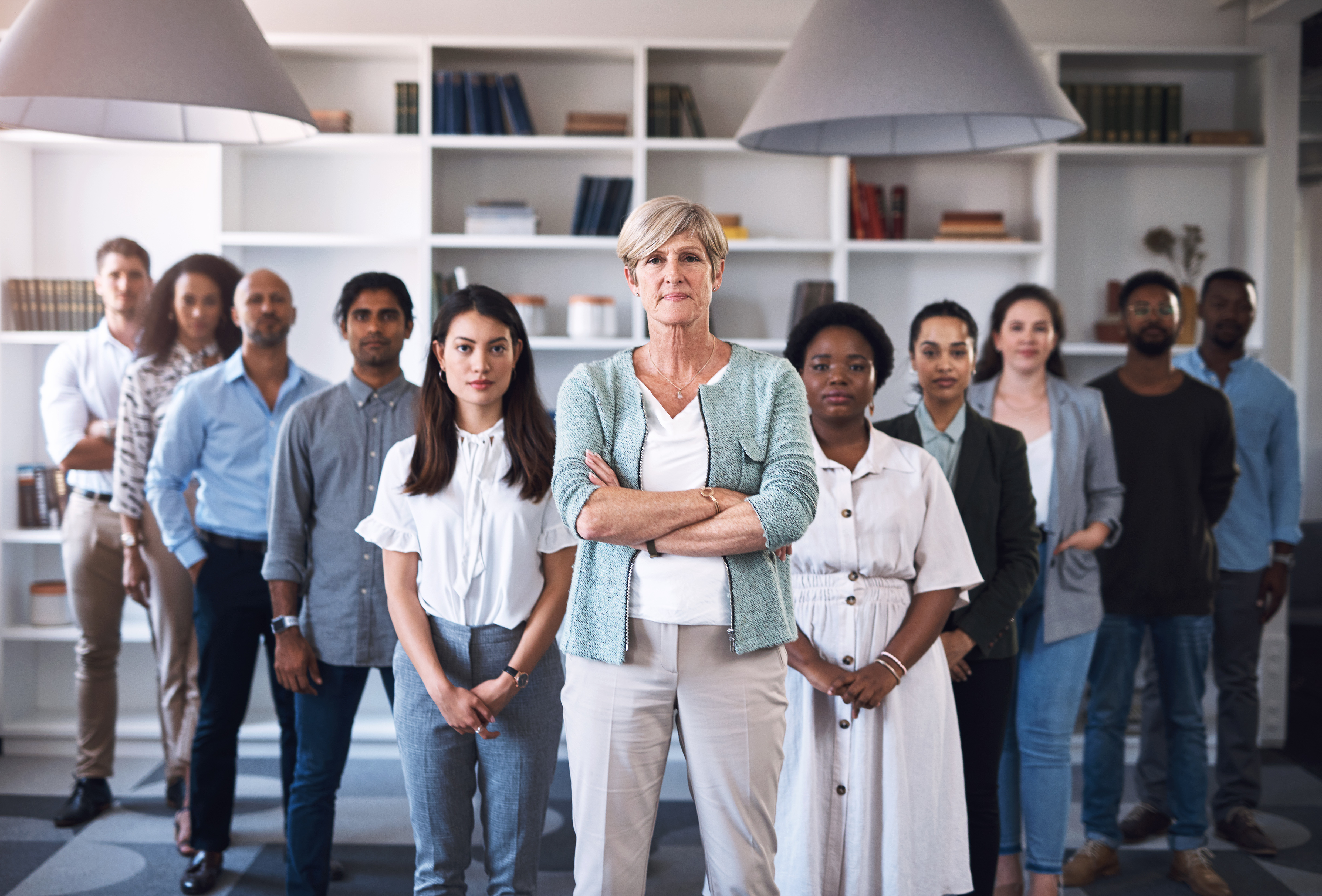 A professional group portrait of a diverse team of eleven staff members in an office with white bookshelves, led by a woman with her arms crossed in the foreground.
