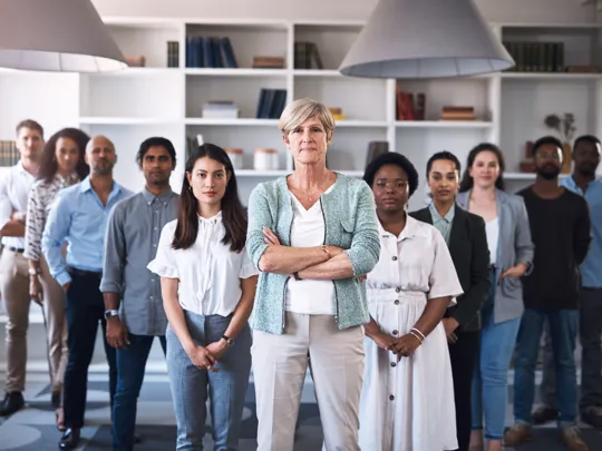 A professional group portrait of a diverse team of eleven staff members in an office with white bookshelves, led by a woman with her arms crossed in the foreground.