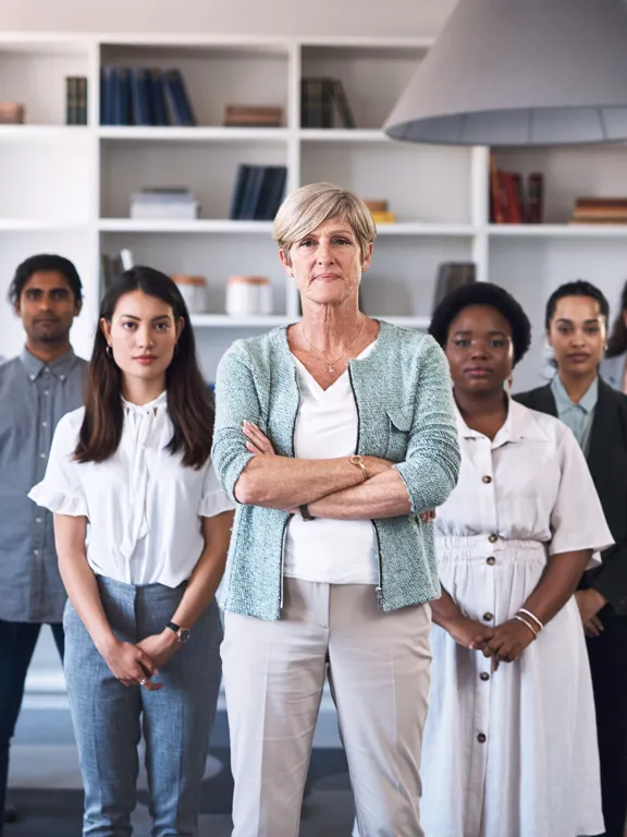 A professional group portrait of a diverse team of eleven staff members in an office with white bookshelves, led by a woman with her arms crossed in the foreground.