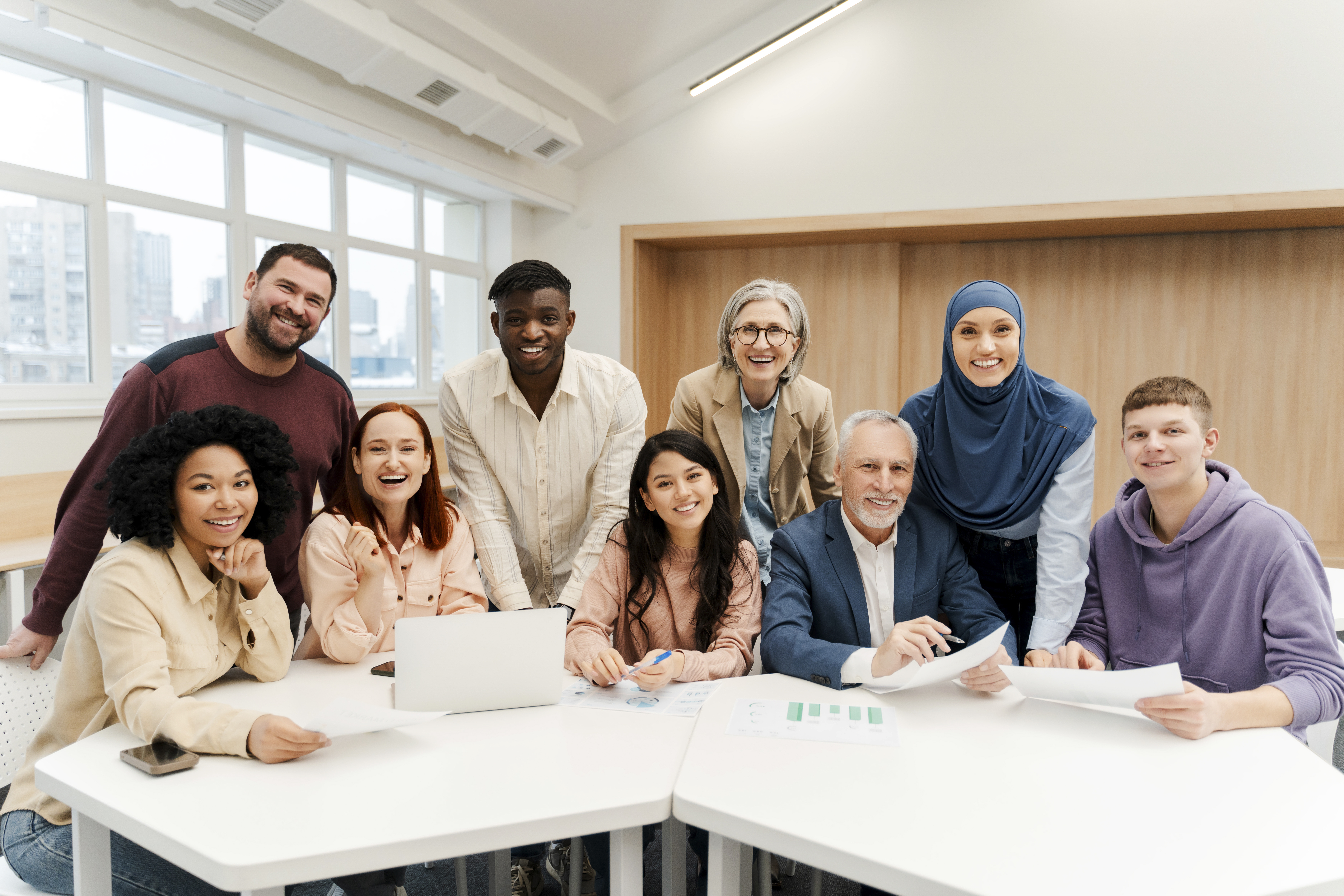 A diverse group of nine smiling professionals, including a woman in a blue hijab and a man with a beard, seated around a conference table in a modern office with a laptop and documents.