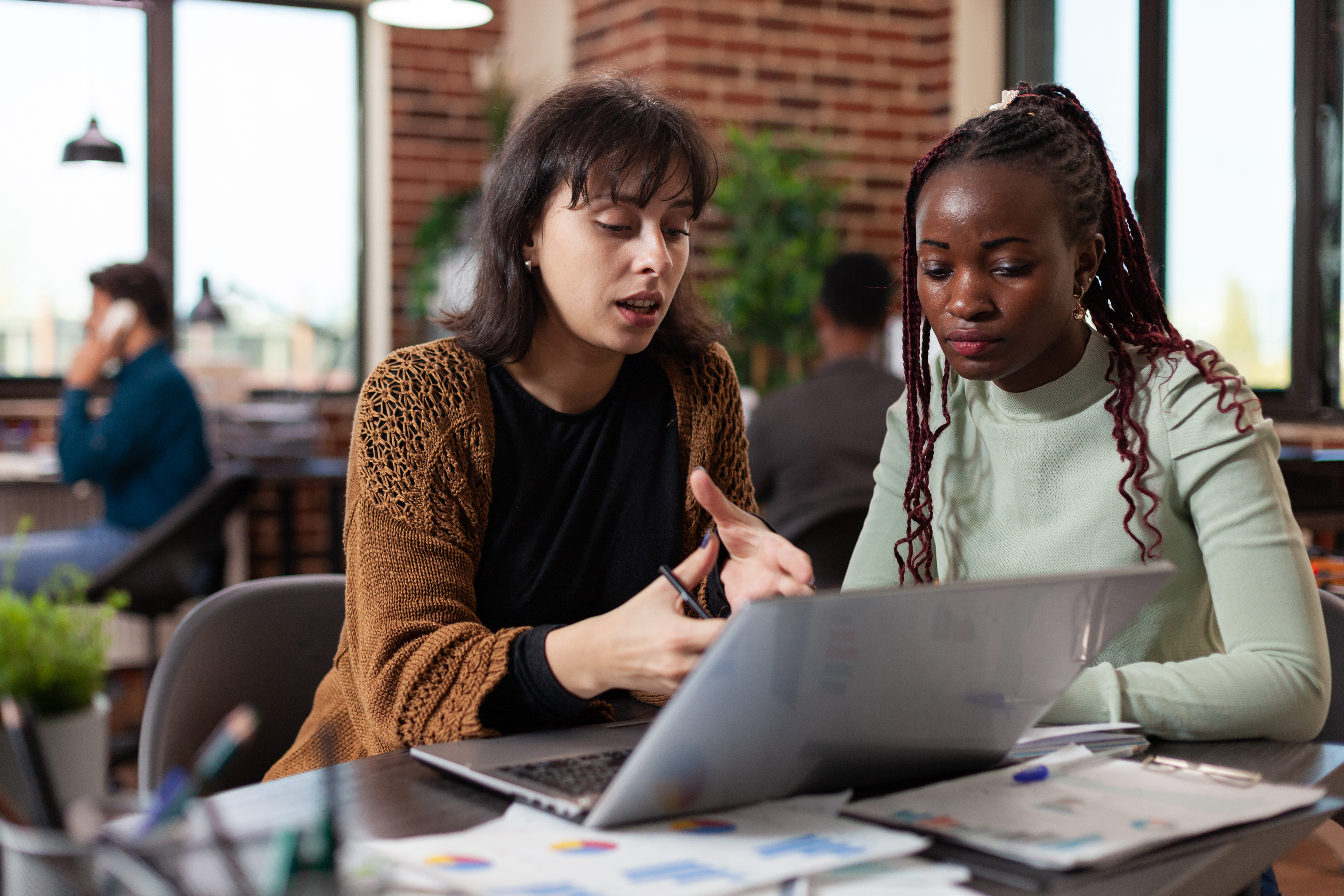 two women sitting at a table working together and discussing content on a laptop in a modern office or study space.