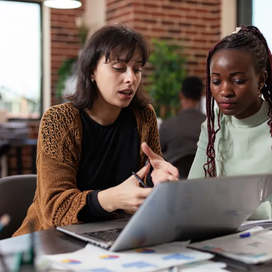 two women sitting at a table working together and discussing content on a laptop in a modern office or study space.