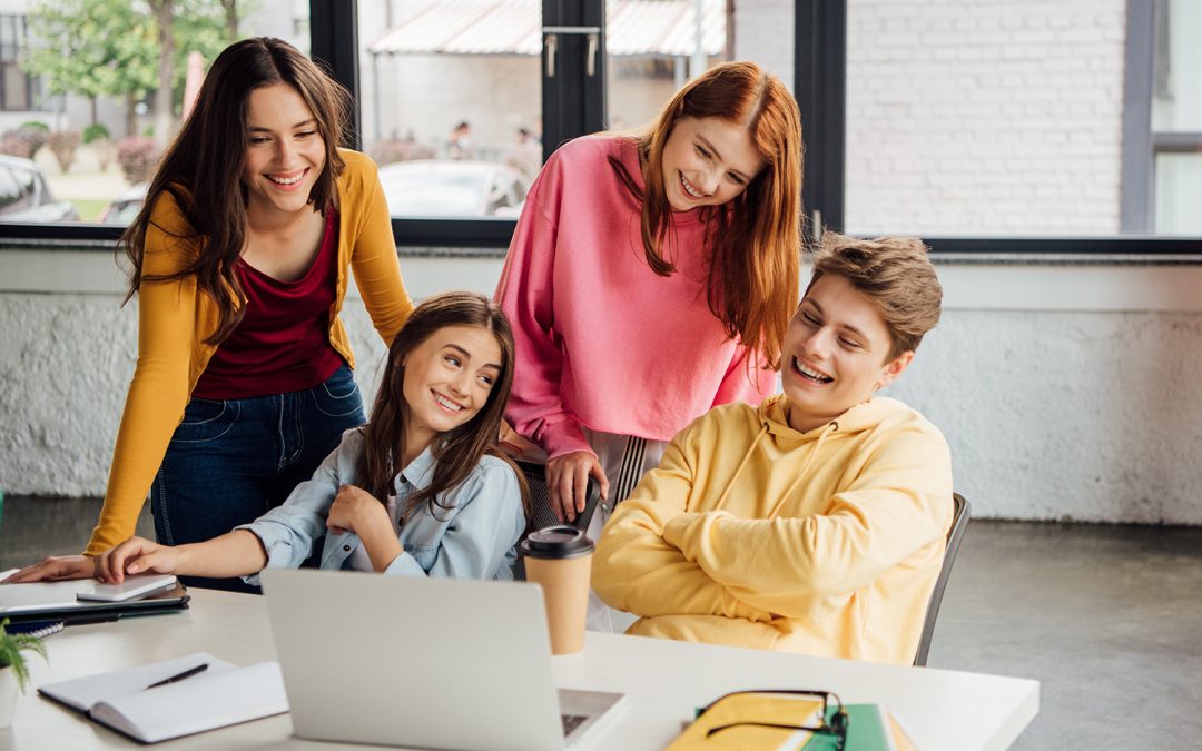 Four happy young students, two females and one male, are gathered around a laptop on a table, laughing and collaborating on a project. They are dressed casually in bright colors, with notebooks and coffee cups nearby, working in a modern, bright study space.
