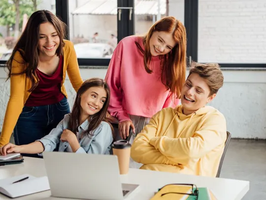 Four happy young students, two females and one male, are gathered around a laptop on a table, laughing and collaborating on a project. They are dressed casually in bright colors, with notebooks and coffee cups nearby, working in a modern, bright study space.