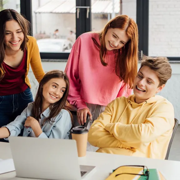Four happy young students, two females and one male, are gathered around a laptop on a table, laughing and collaborating on a project. They are dressed casually in bright colors, with notebooks and coffee cups nearby, working in a modern, bright study space.